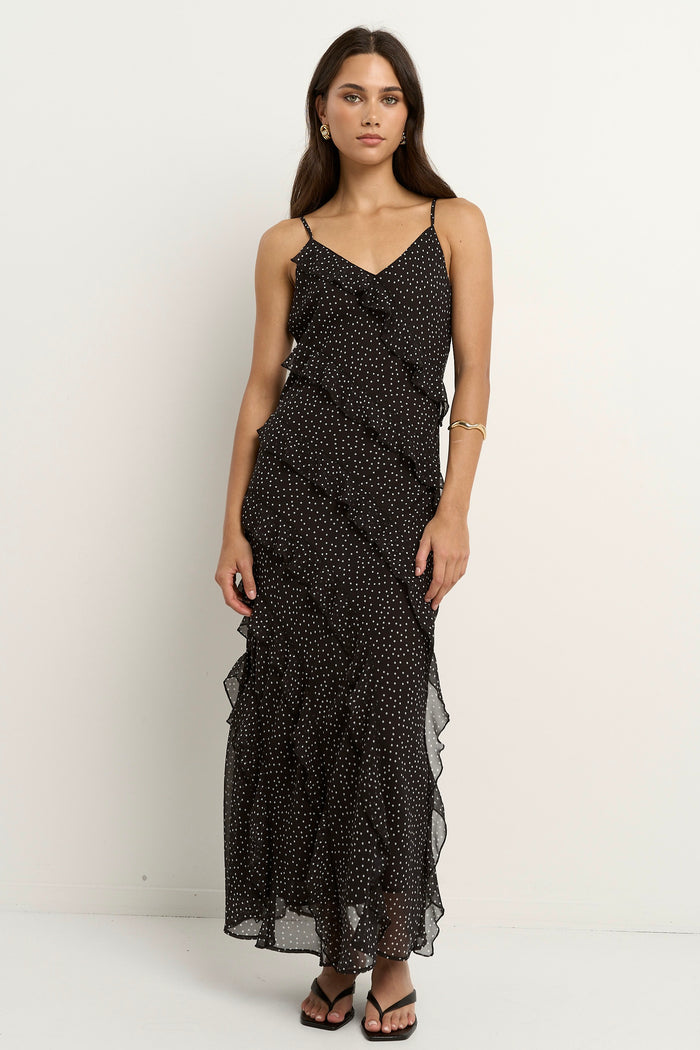 Woman wearing a black bias-cut polka dot dress with delicate straps and cascading ruffles, standing against a neutral wall.