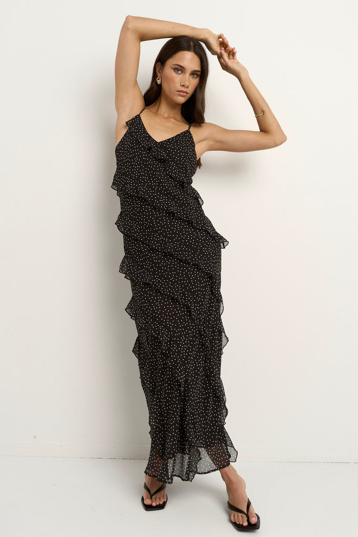 Woman wearing a black bias-cut polka dot dress with delicate straps and cascading ruffles, standing against a neutral wall.