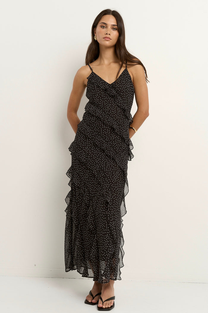 Woman wearing a black bias-cut polka dot dress with delicate straps and cascading ruffles, standing against a neutral wall.
