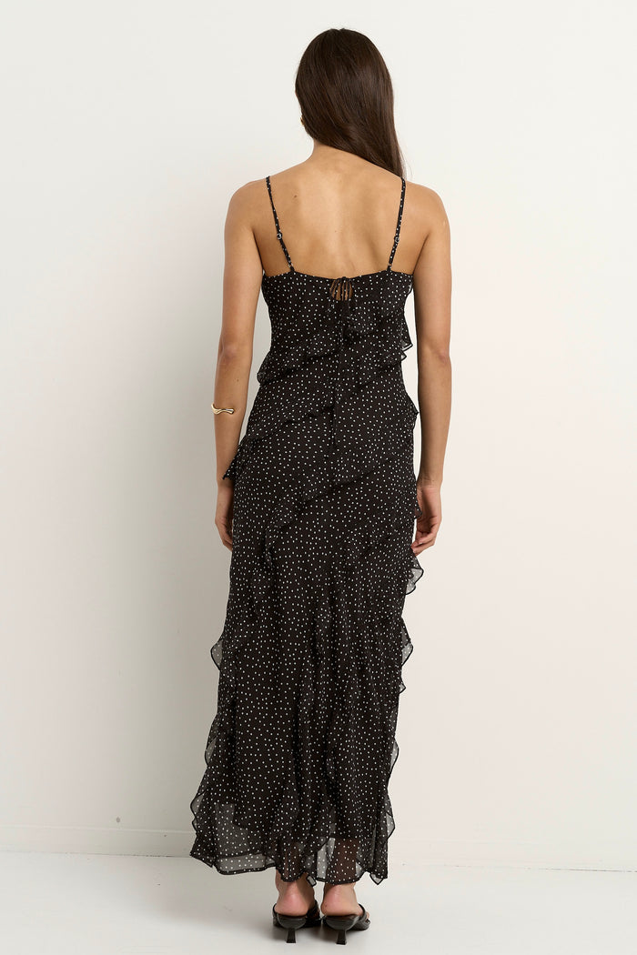 Woman wearing a black bias-cut polka dot dress with delicate straps and cascading ruffles, standing against a neutral wall.