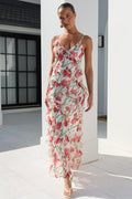 A woman models a sleeveless, ruffled maxi dress with a red and pink floral print on a white background, walking in a bright, modern white outdoor corridor.