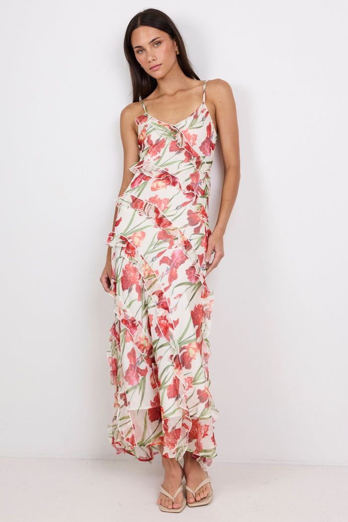 A woman modeling a sleeveless maxi dress with thin straps and a red, pink, and green floral print on a light background, standing against a plain white studio backdrop.