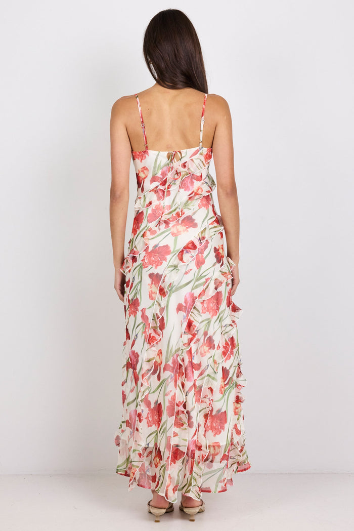 A woman viewed from behind wearing a sleeveless maxi dress with thin straps and a red, pink, and green floral print on a light background, standing against a plain white studio backdrop.
