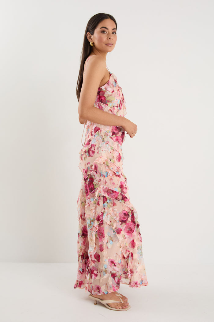 Woman wearing a strappy floral bias-cut maxi dress in pink and pastel tones with cascading ruffle layers, styled with neutral sandals.