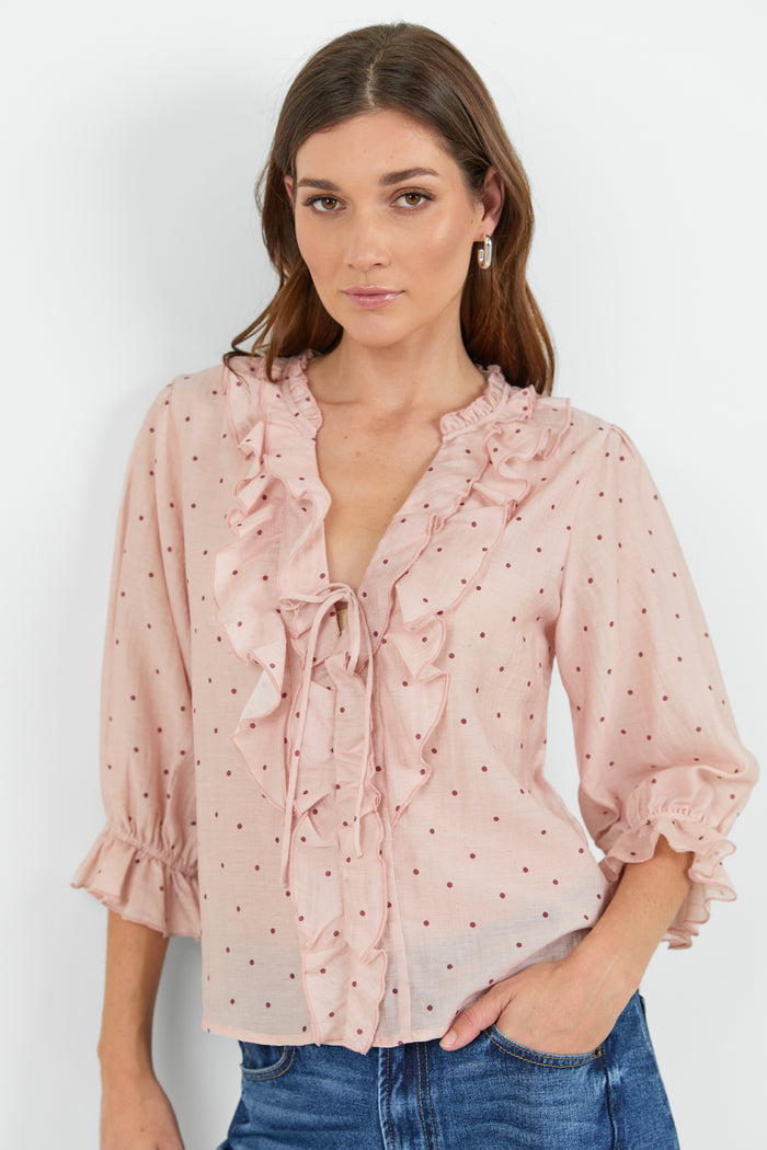A woman wearing a light pink ruffled blouse with small dark polka dots and blue jeans, standing against a plain light gray studio background.