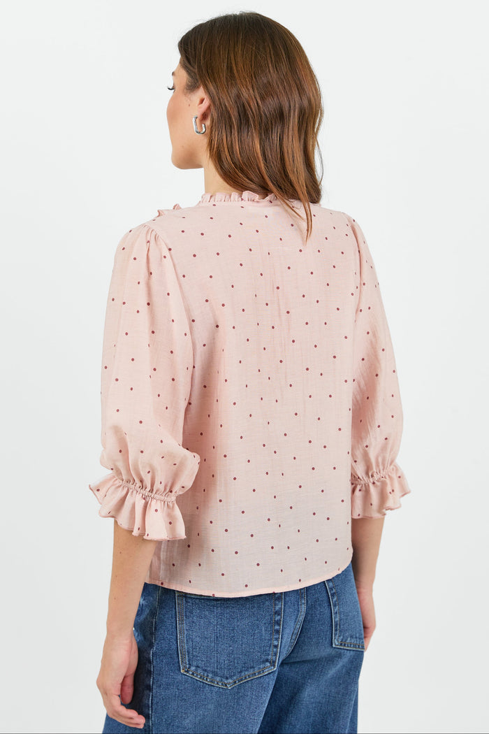 A woman shown from the back wearing a light pink blouse with small dark polka dots, ruffled trim at the collar and cuffs, paired with blue jeans against a plain white studio background.
col_pattern