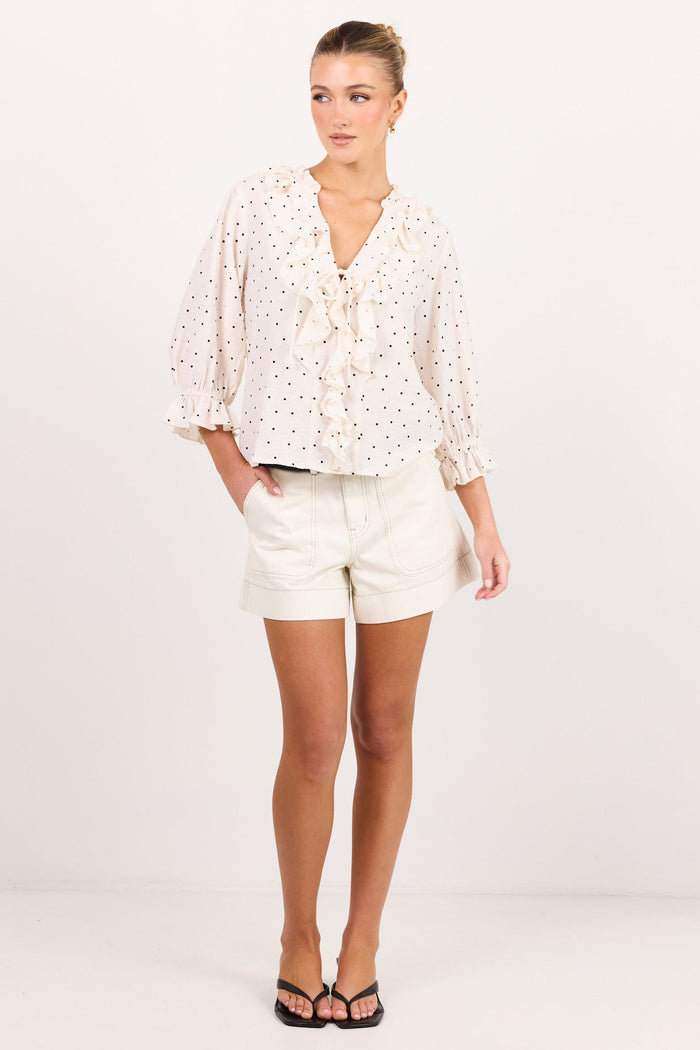 A woman modeling a white eyelet blouse with small black polka dots and a tie neckline, paired with white shorts and black sandals, standing against a plain white studio background.