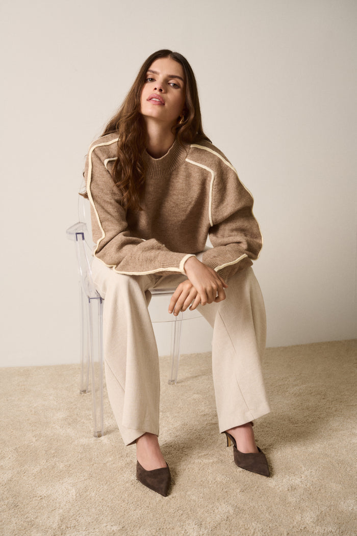 Woman sitting on a chair wearing a brown sweater and beige pants against a plain background