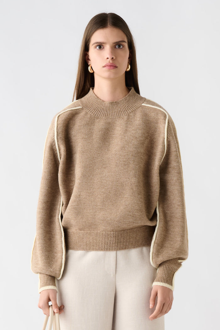 Woman wearing a beige sweater and white pants, holding a beige handbag on a white background