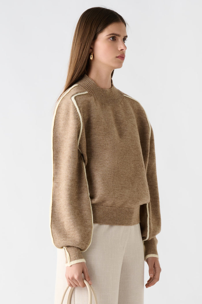 Woman wearing a beige sweater and white pants holding a cream handbag on a light gray background