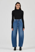 A model stands against a light gray studio background wearing a black long-sleeve mock-neck top, high-waisted wide-leg blue jeans, and black ankle boots.