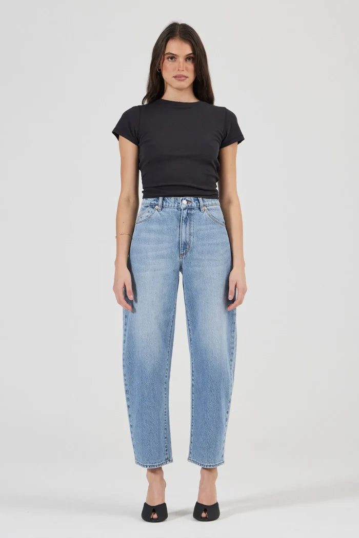 A woman modeling a pair of light-wash blue cropped jeans with a black short-sleeve t-shirt and black flat shoes, standing against a light gray studio background.