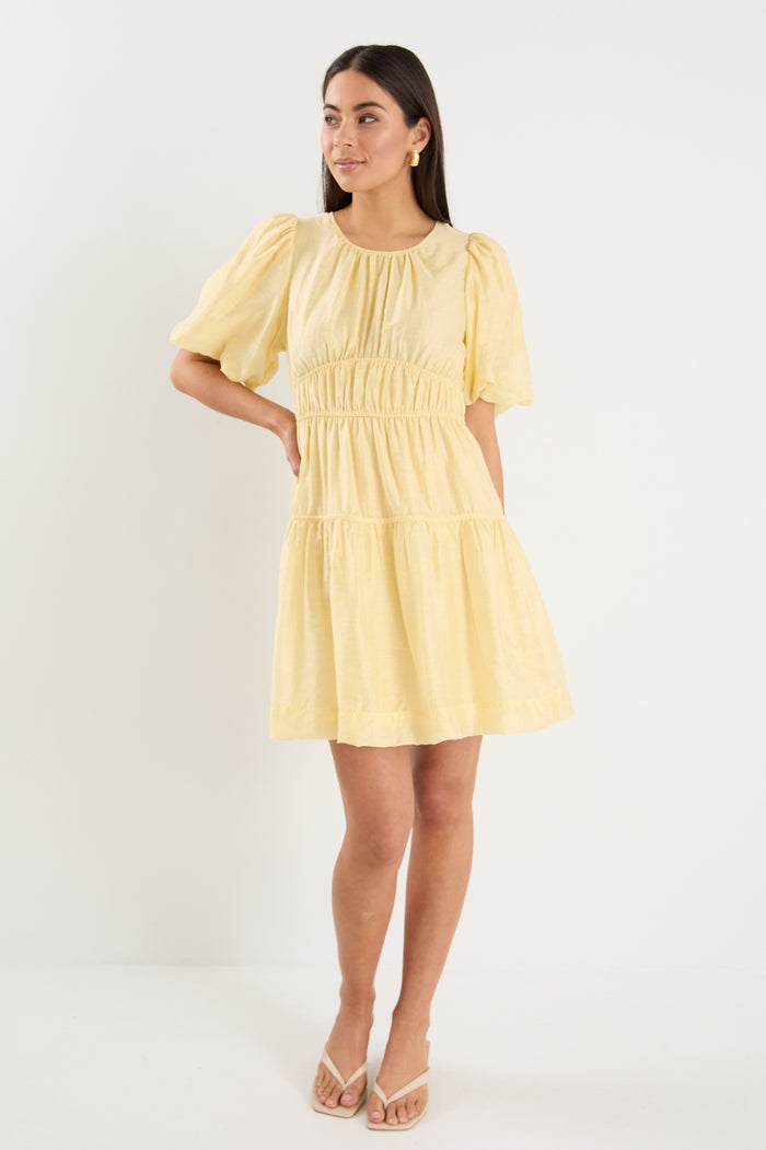 Woman wearing a short butter-yellow mini dress with puff sleeves, elastic waist, and back tie detail, styled with neutral sandals.