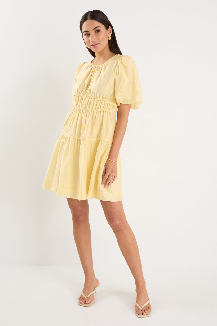 Woman wearing a short butter-yellow mini dress with puff sleeves, elastic waist, and back tie detail, styled with neutral sandals.