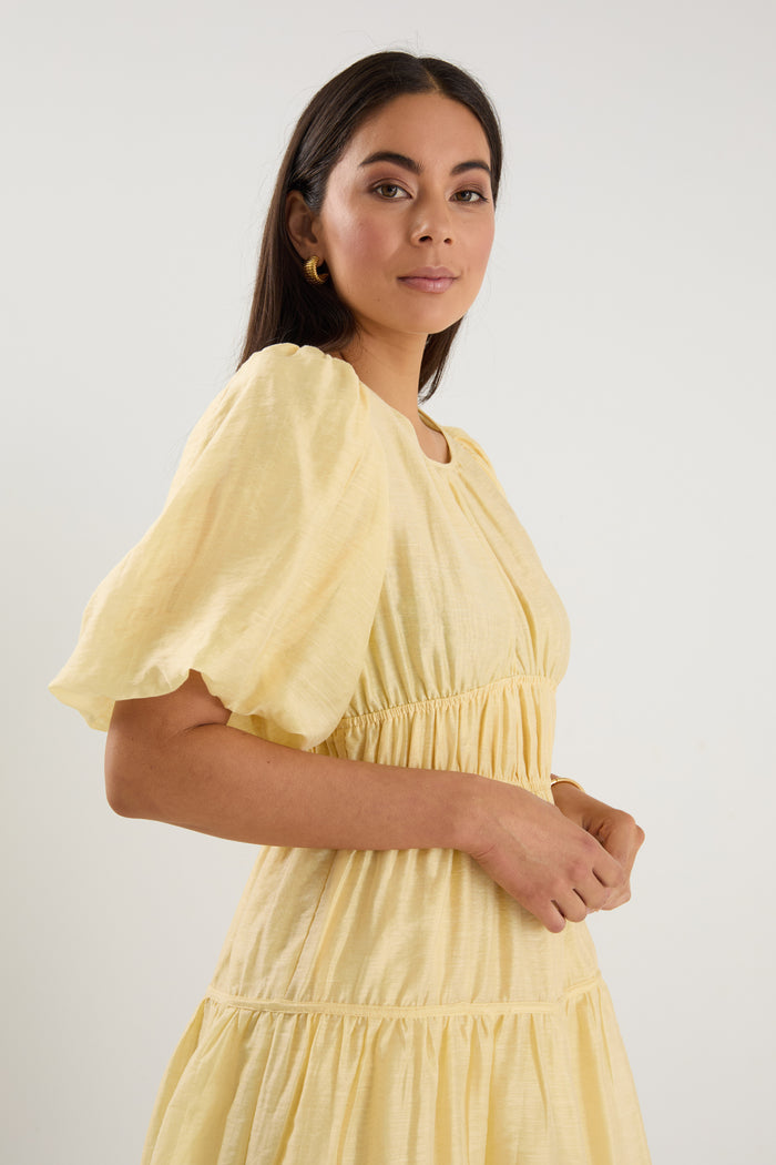 Woman wearing a short butter-yellow mini dress with puff sleeves, elastic waist, and back tie detail, styled with neutral sandals.