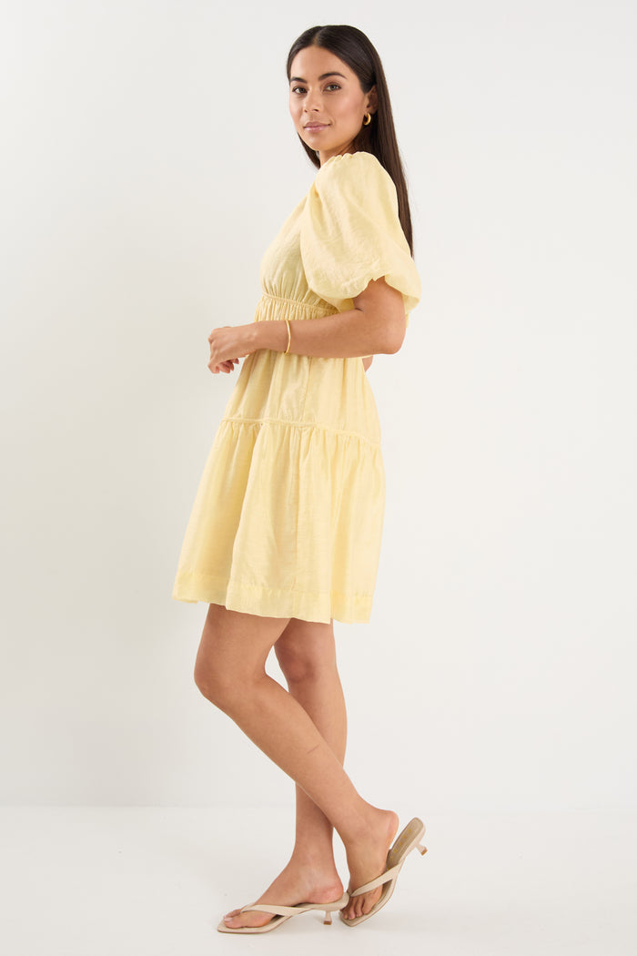 Woman wearing a short butter-yellow mini dress with puff sleeves, elastic waist, and back tie detail, styled with neutral sandals.