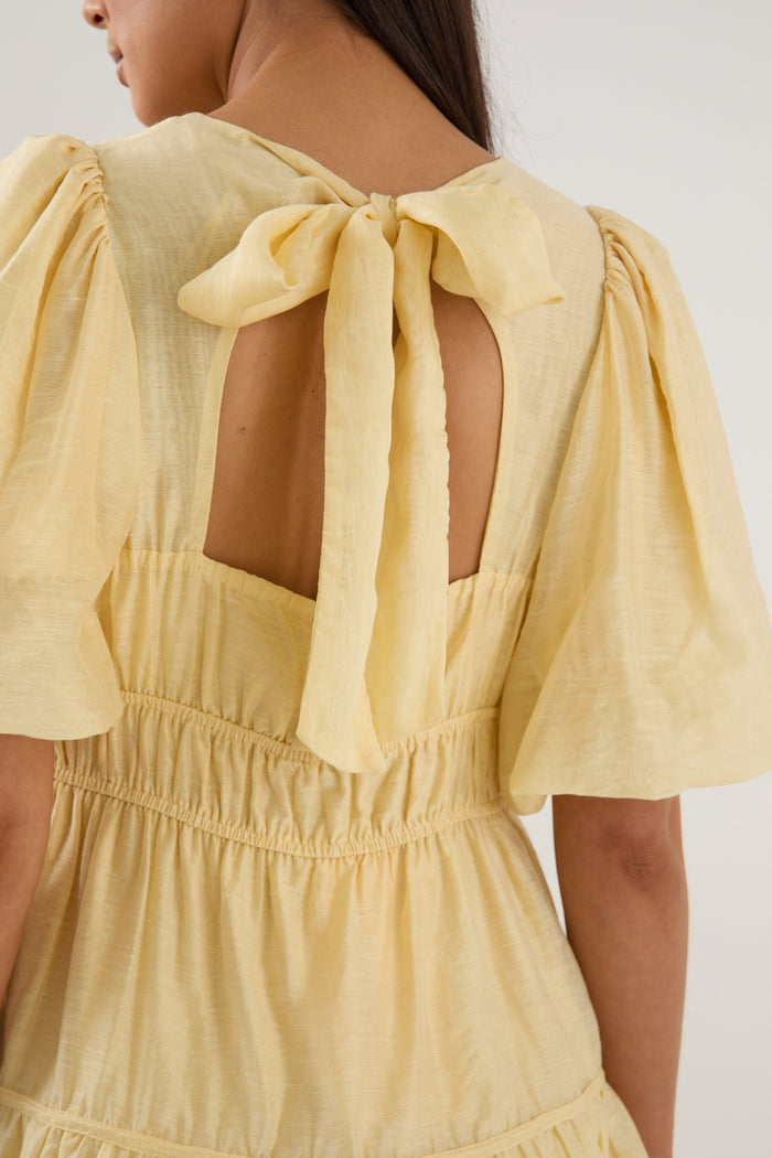 Woman wearing a short butter-yellow mini dress with puff sleeves, elastic waist, and back tie detail, styled with neutral sandals.