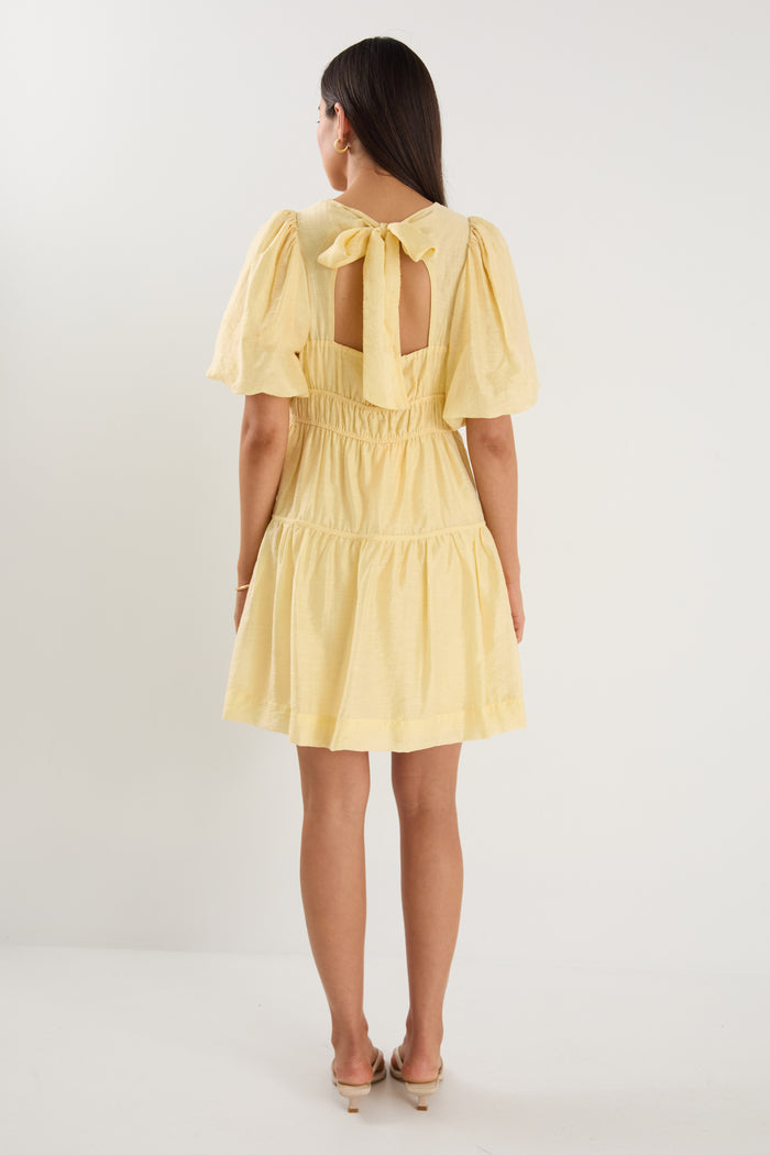 Woman wearing a short butter-yellow mini dress with puff sleeves, elastic waist, and back tie detail, styled with neutral sandals.
