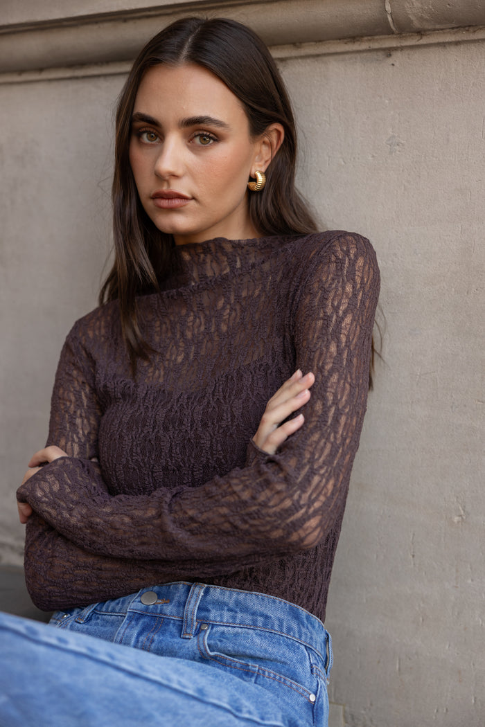 Woman wearing a dark lace top and blue jeans against a neutral wall.