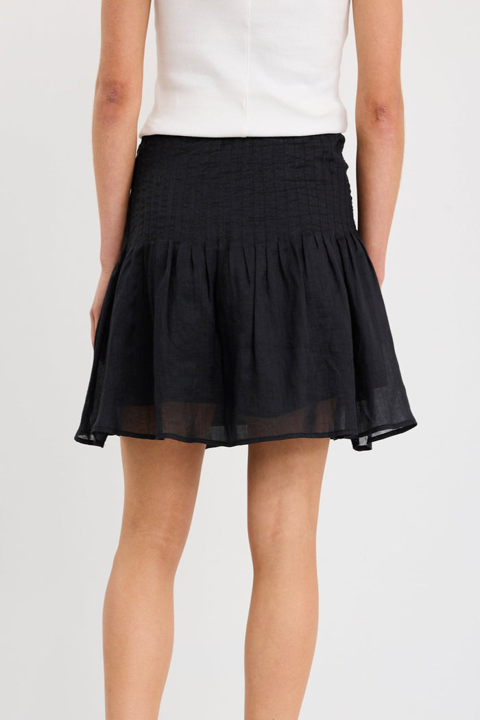Woman wearing a black mini skirt with pintuck waist detail and soft flared shape, styled with a white tank and black sandals.