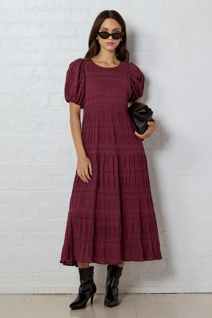 Woman wearing a burgundy dress with puff sleeves against a white brick wall.