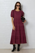 Woman wearing a burgundy dress with puff sleeves against a white brick wall.