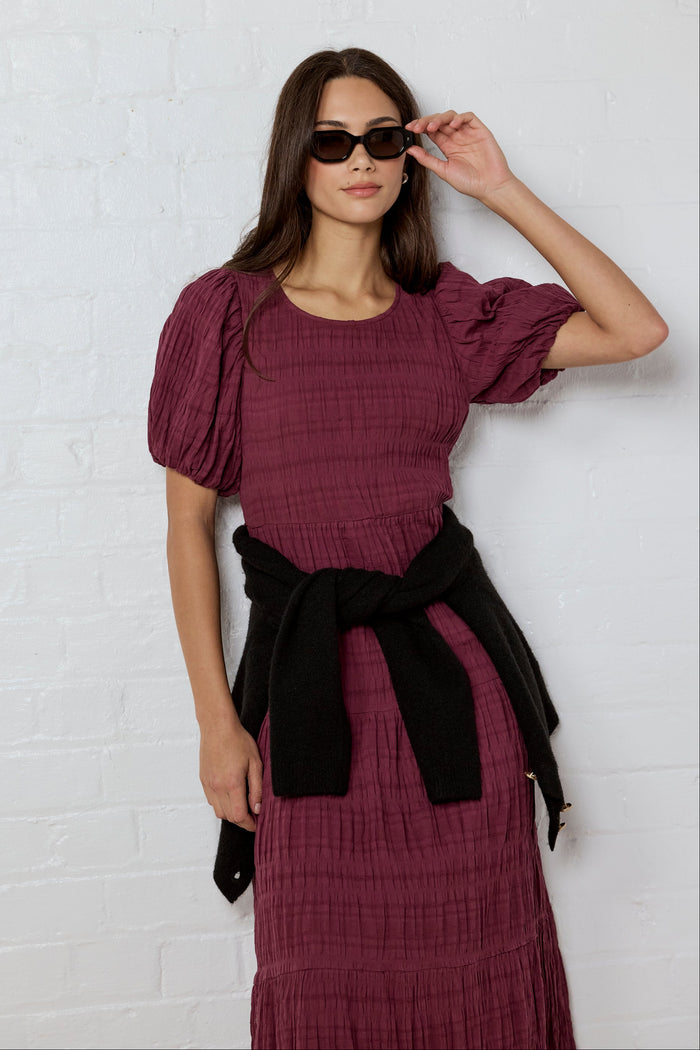 Woman wearing a burgundy dress with puffed sleeves against a white brick wall.