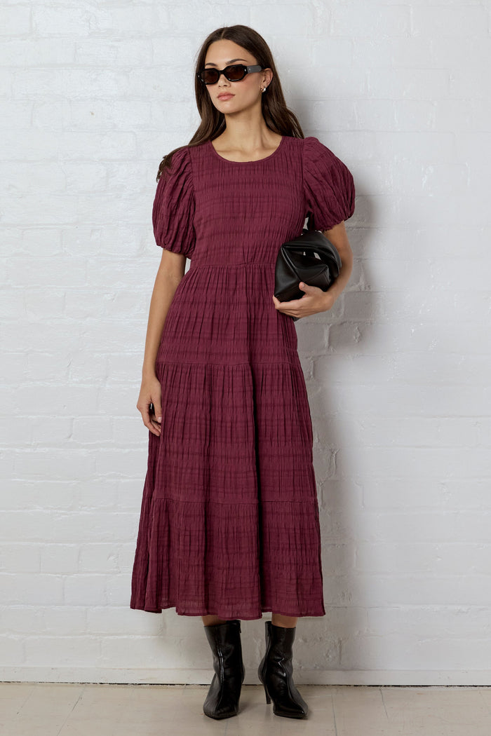 Woman wearing a burgundy dress with puff sleeves against a white brick wall.