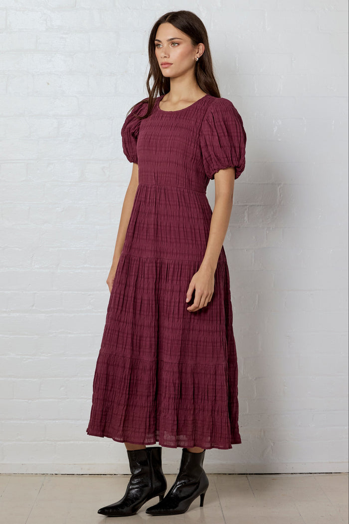Woman wearing a burgundy dress with puff sleeves against a white brick wall.