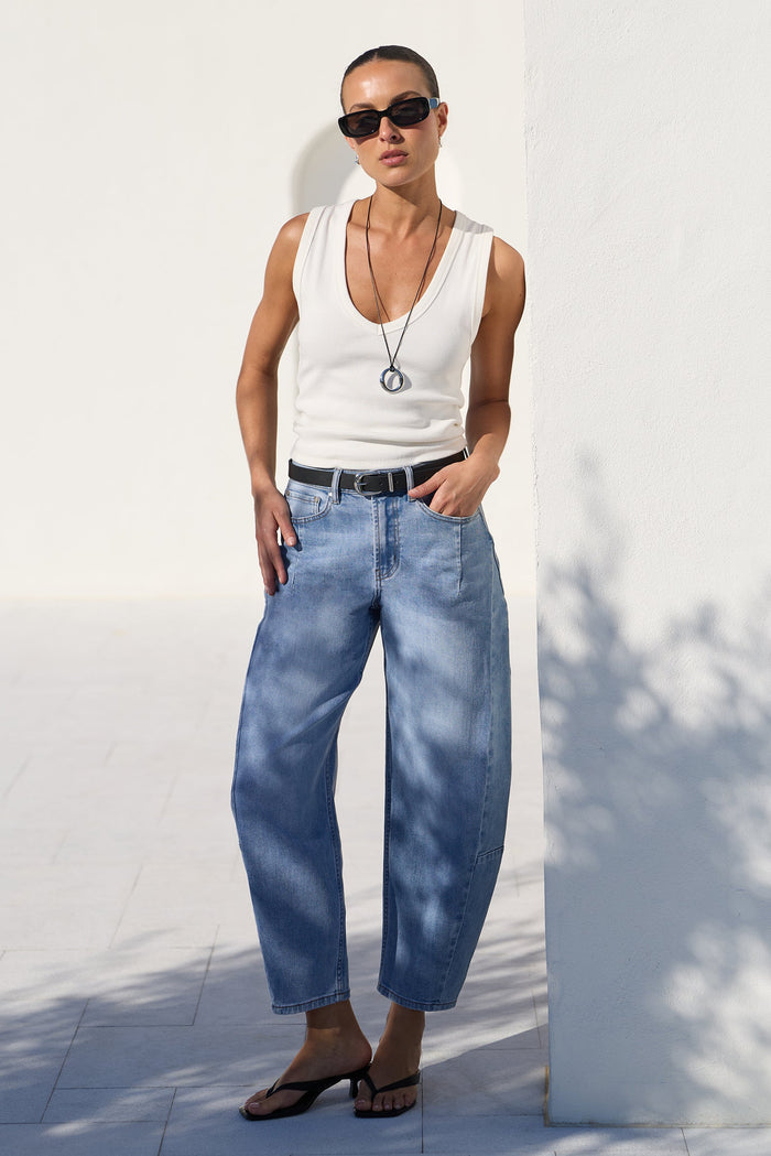 A model wearing a white tank top, wide-leg light blue jeans, a black belt, and black sandals, standing outdoors against a white wall.