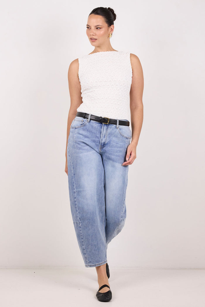 A woman modeling a pair of light-wash, wide-leg cropped jeans with a black belt, paired with a white sleeveless top and black flats, against a plain white studio background.