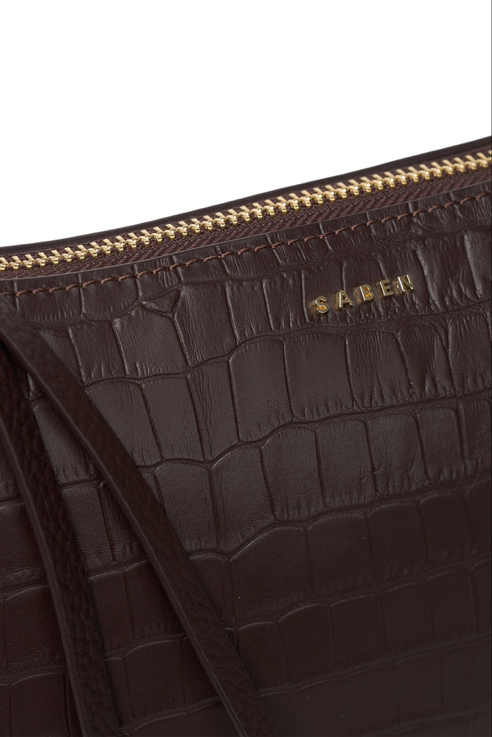 Close-up of a dark brown crocodile-embossed leather bag with a gold zipper and gold SABEN logo on a white background.