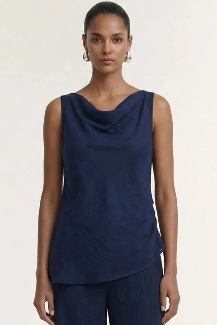 A model wearing a sleeveless navy blue cowl-neck top and matching navy pleated trousers, photographed against a plain off-white studio background.