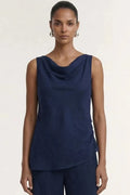A model wearing a sleeveless navy blue cowl-neck top and matching navy pleated trousers, photographed against a plain off-white studio background.