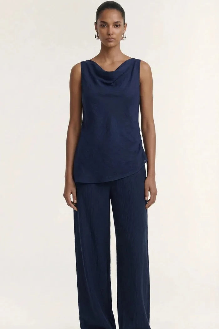 A model stands against a plain light beige studio background wearing a dark navy sleeveless draped top and matching wide-leg pants, with brown sandals.