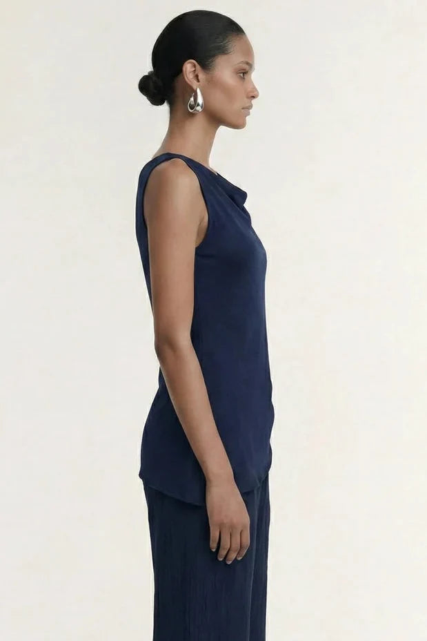 A model is shown in profile wearing a matching navy blue sleeveless top and pleated wide-leg trousers set, styled with simple brown sandals against a clean, off-white studio background.