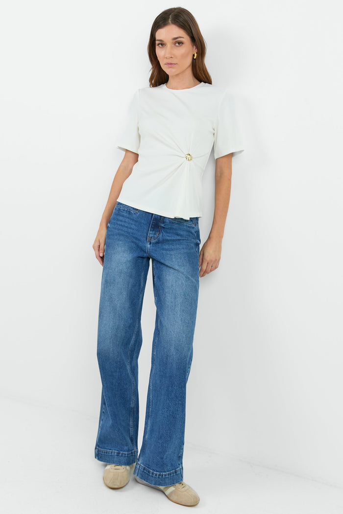 Woman wearing a white t-shirt with a twist and clasp detail and blue jeans on a white background