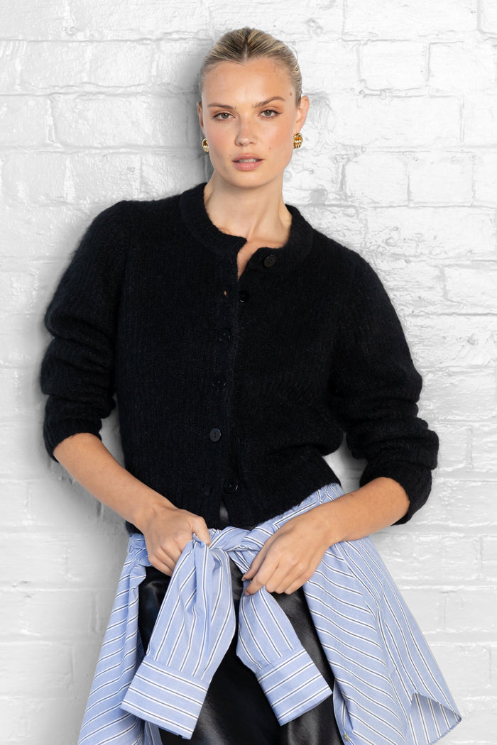 Woman wearing a black cardigan over a blue shirt against a white brick wall