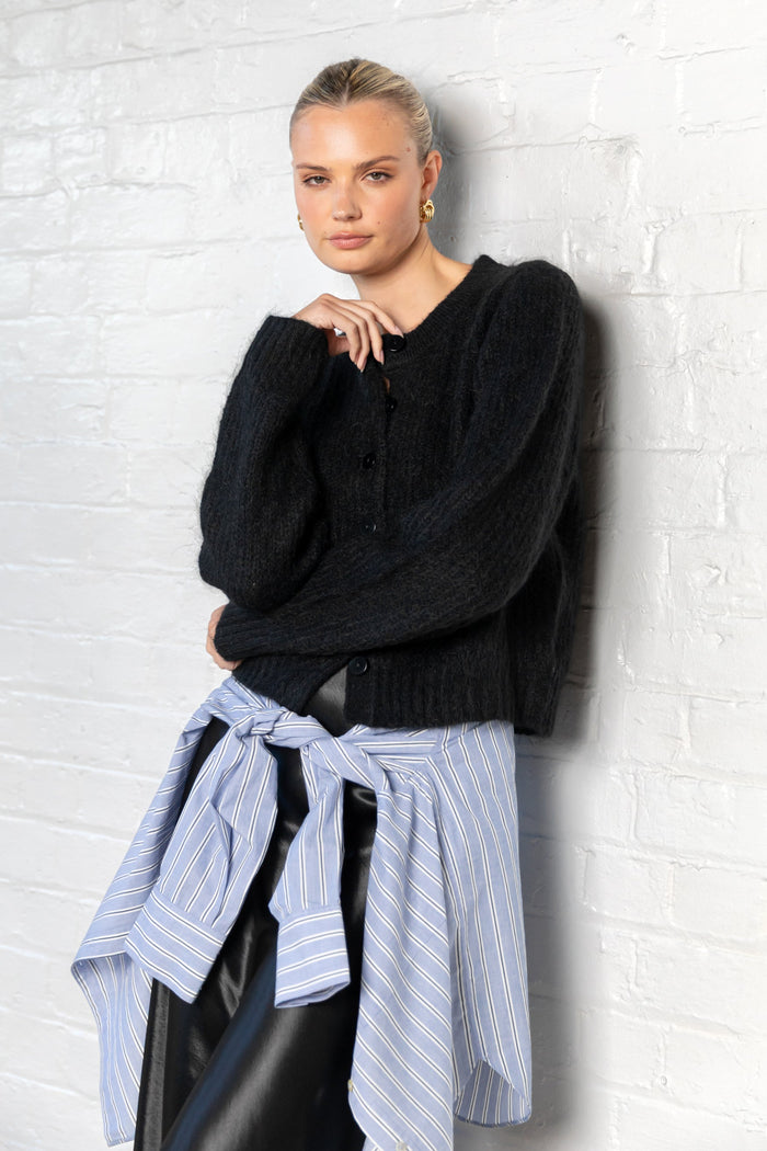 Woman wearing a black sweater and striped shirt against a white brick wall
