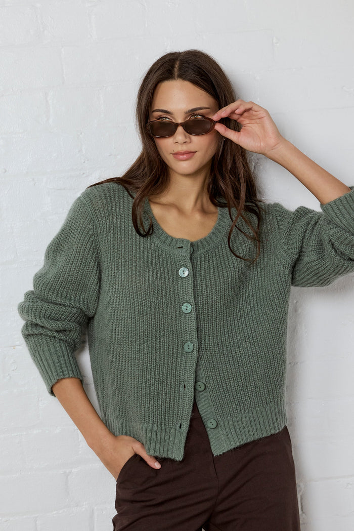Woman wearing a green knitted cardigan and sunglasses against a white brick wall.