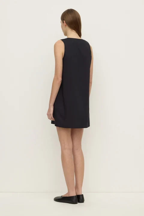 A woman stands with her back to the camera wearing a sleeveless black shift dress and black flats against a plain white studio background.