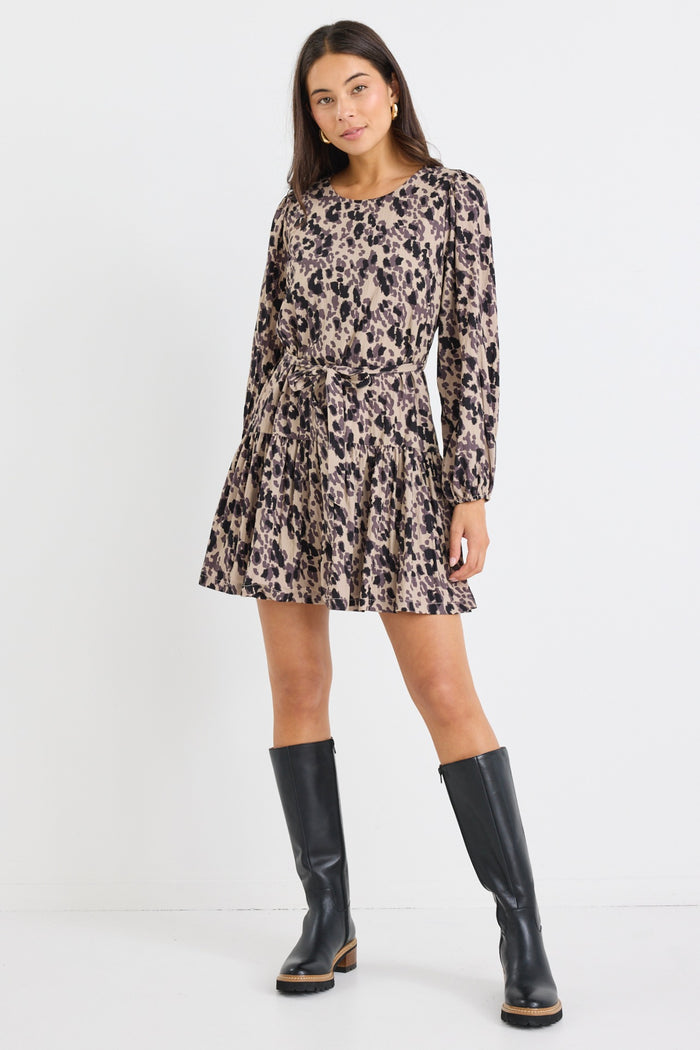Woman wearing a long-sleeve mini dress with an animal print pattern, belted at the waist with a flared tiered skirt and black boots.