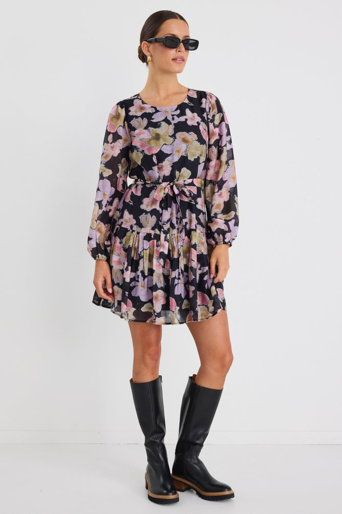 Woman wearing a long-sleeve floral mini dress with a tie waist and tiered skirt, styled with knee-high black boots and sunglasses.