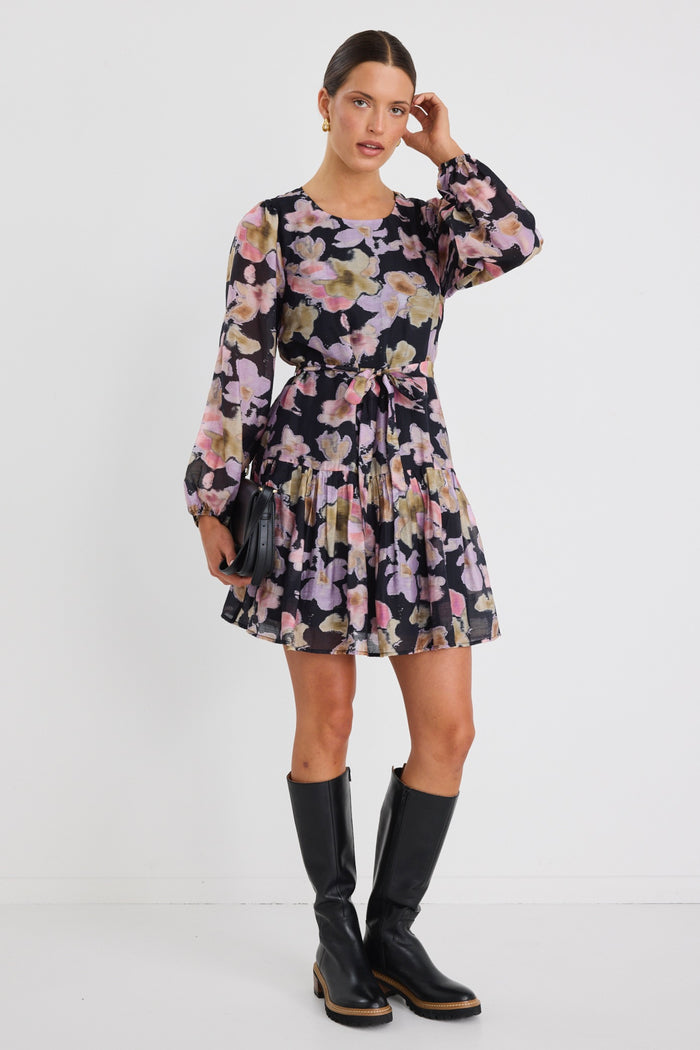 Woman wearing a long-sleeve floral mini dress with a tie waist and tiered skirt, styled with knee-high black boots and sunglasses.
