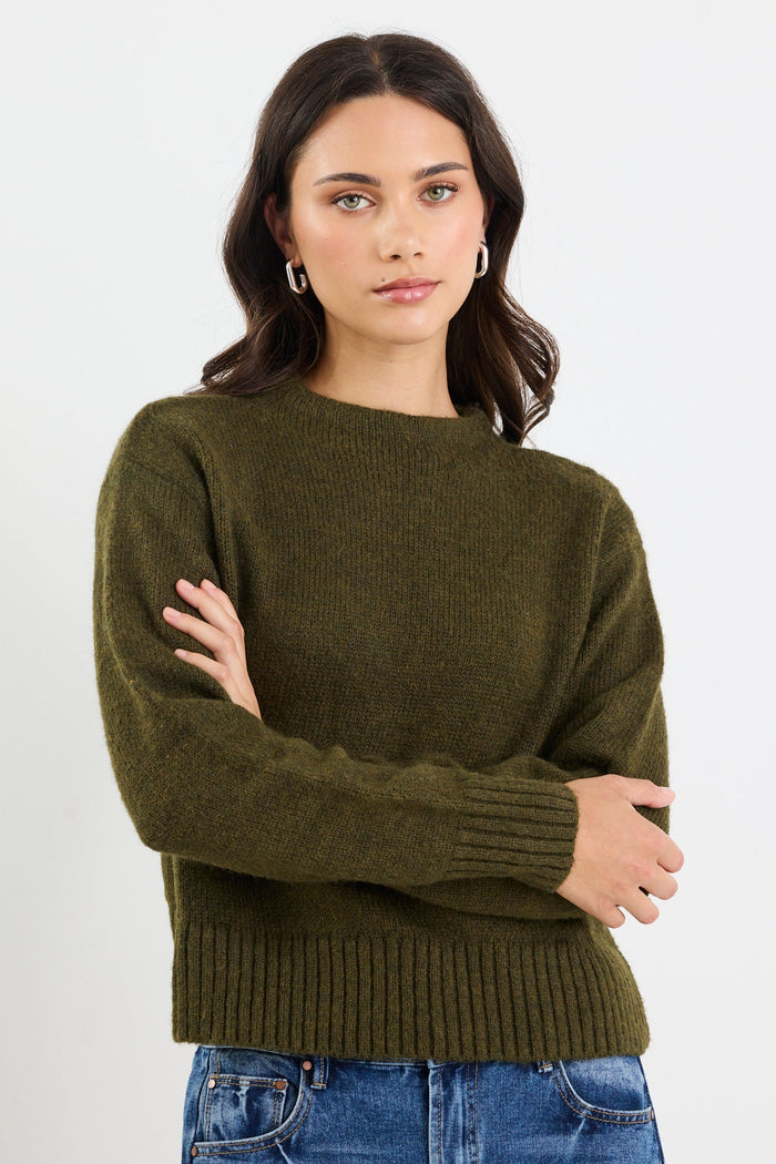 Woman wearing an olive green sweater and blue jeans against a white background