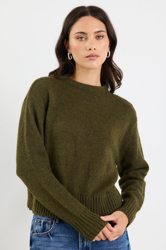 Woman wearing an olive green sweater and blue jeans on a white background