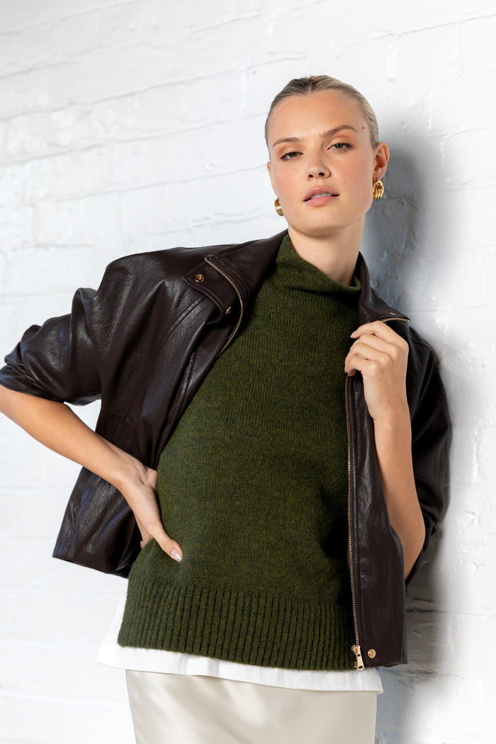 Woman wearing a green sweater and black leather jacket against a white brick wall.