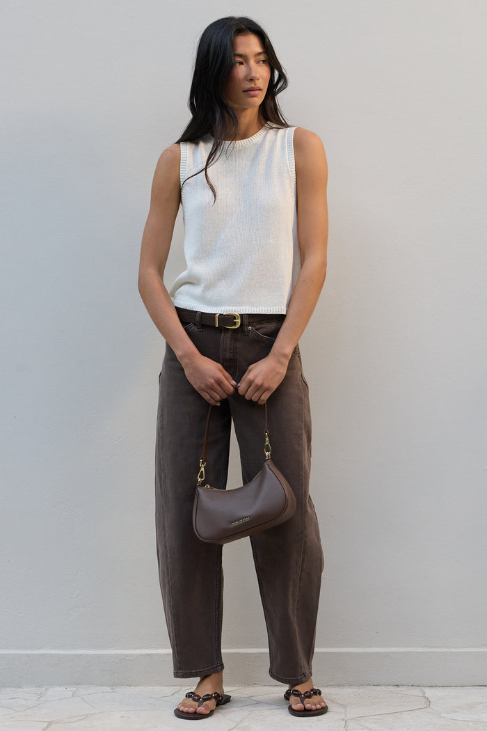 A woman stands against a light gray wall wearing a sleeveless white ribbed knit top, dark brown wide-leg pants, and brown sandals, while holding a taupe crescent-shaped shoulder bag.