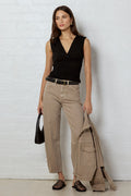 A woman stands against a white brick wall wearing a black sleeveless wrap top, beige wide-leg cargo pants, and black patterned sandals, while holding a black handbag and a beige jacket.