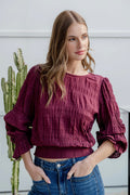 Woman wearing a burgundy long sleeve top with shirred detailing and fluted cuffs, styled with high-waisted blue jeans.